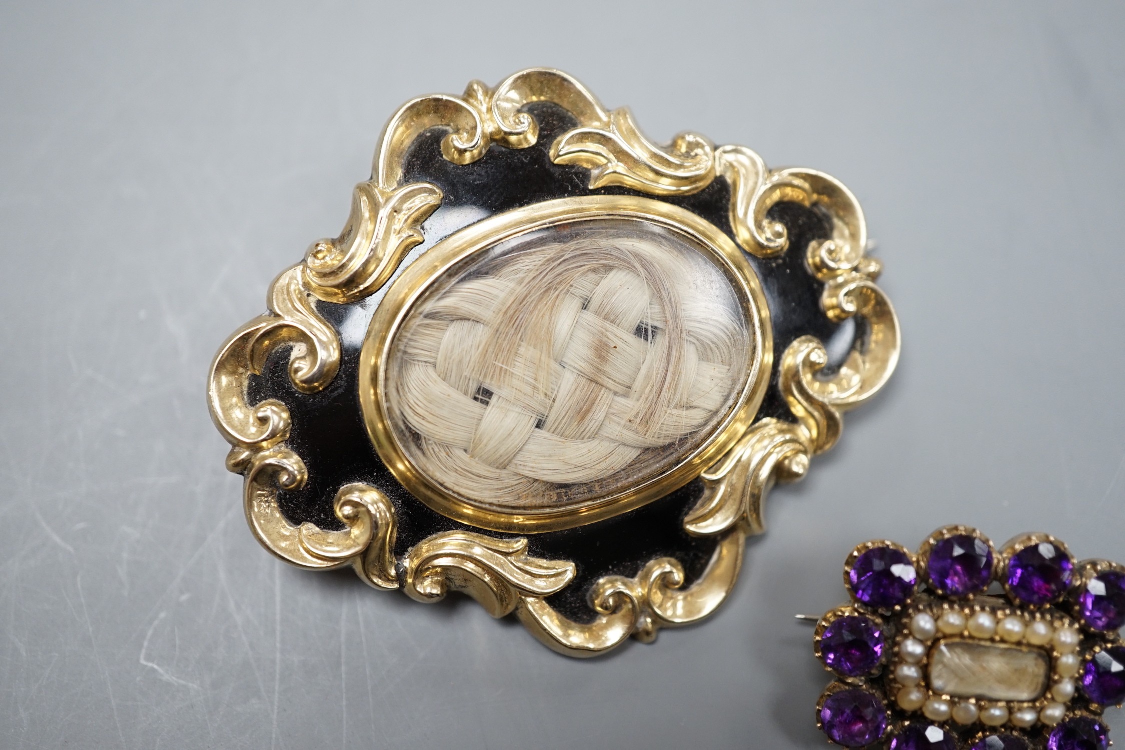 Two Victorian yellow metal (tests as 9ct) mourning brooches, one with black enamel and pendant bale, 51mm, the other with amethyst and seed pearl, both with plaited hair beneath a glazed panel.